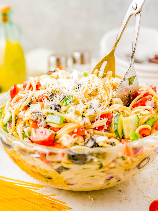 A vibrant bowl of pasta salad with tomatoes, olives, and grated cheese, served with tongs on a kitchen counter.
