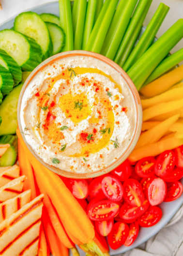 A bowl of hummus surrounded by assorted colorful vegetables like cucumber, celery, tomatoes, and carrots.