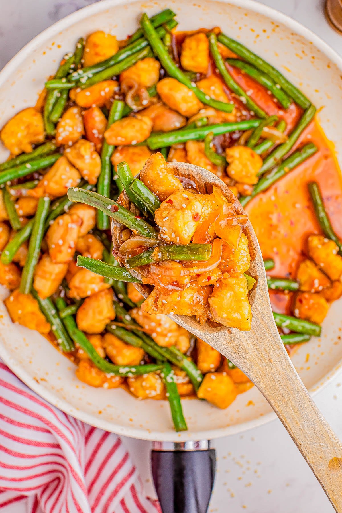 A wooden spoon holds a serving of stir-fried chicken and green beans coated in a glossy sauce, over a pan with more of the meal visible.