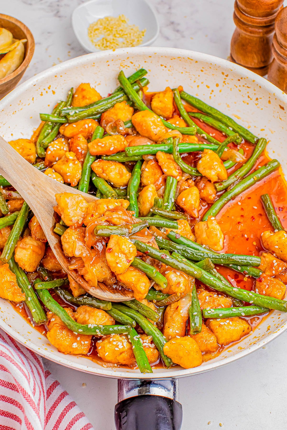 A skillet filled with stir-fried green beans and chunks of chicken in a glossy orange sauce, with a wooden spoon.