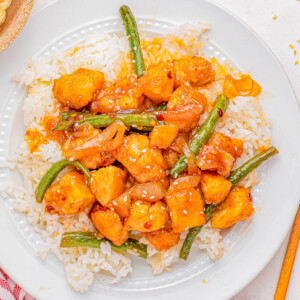 A plate of chicken stir fry with green beans and sesame seeds over white rice, accompanied by chopsticks and a striped napkin.