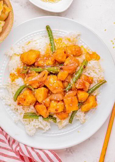 A plate of chicken stir fry with green beans and sesame seeds over white rice, accompanied by chopsticks and a striped napkin.