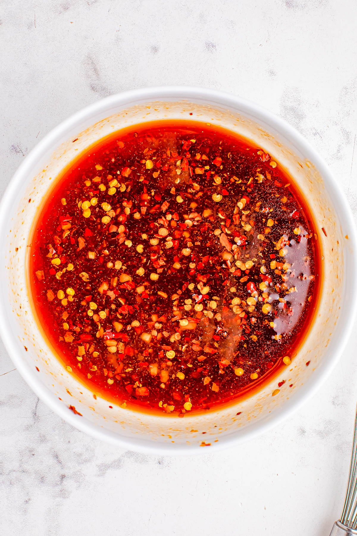 A bowl of chili sauce with floating chili flakes and seeds on a white surface.