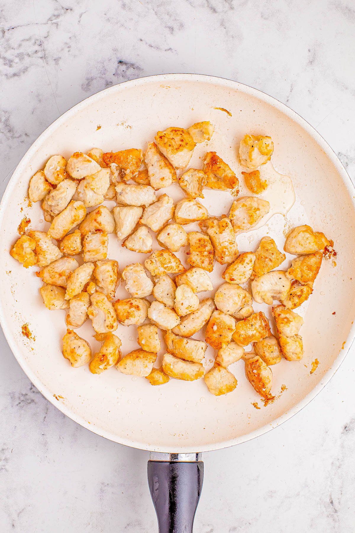 Cooked chicken pieces in a white frying pan on a marble surface.