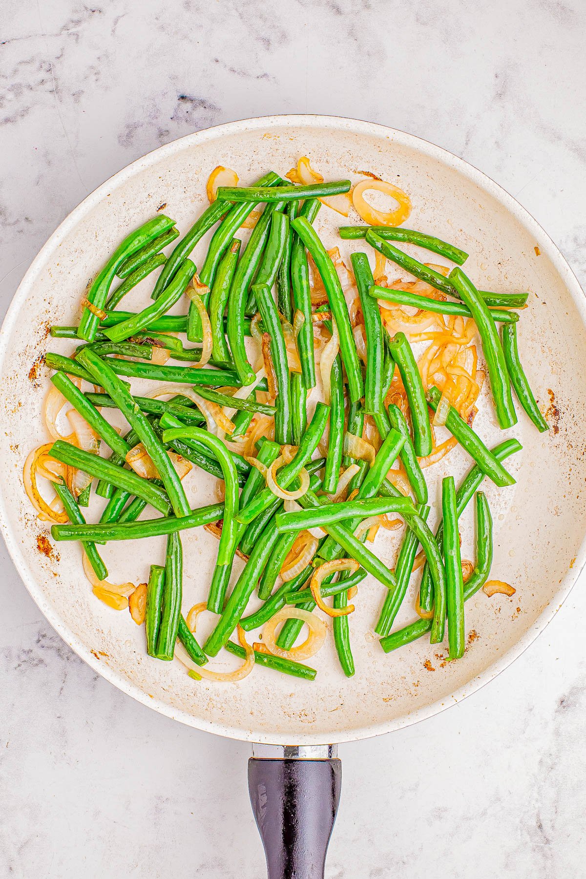 Cooked green beans and sliced onions in a white frying pan on a marble surface.