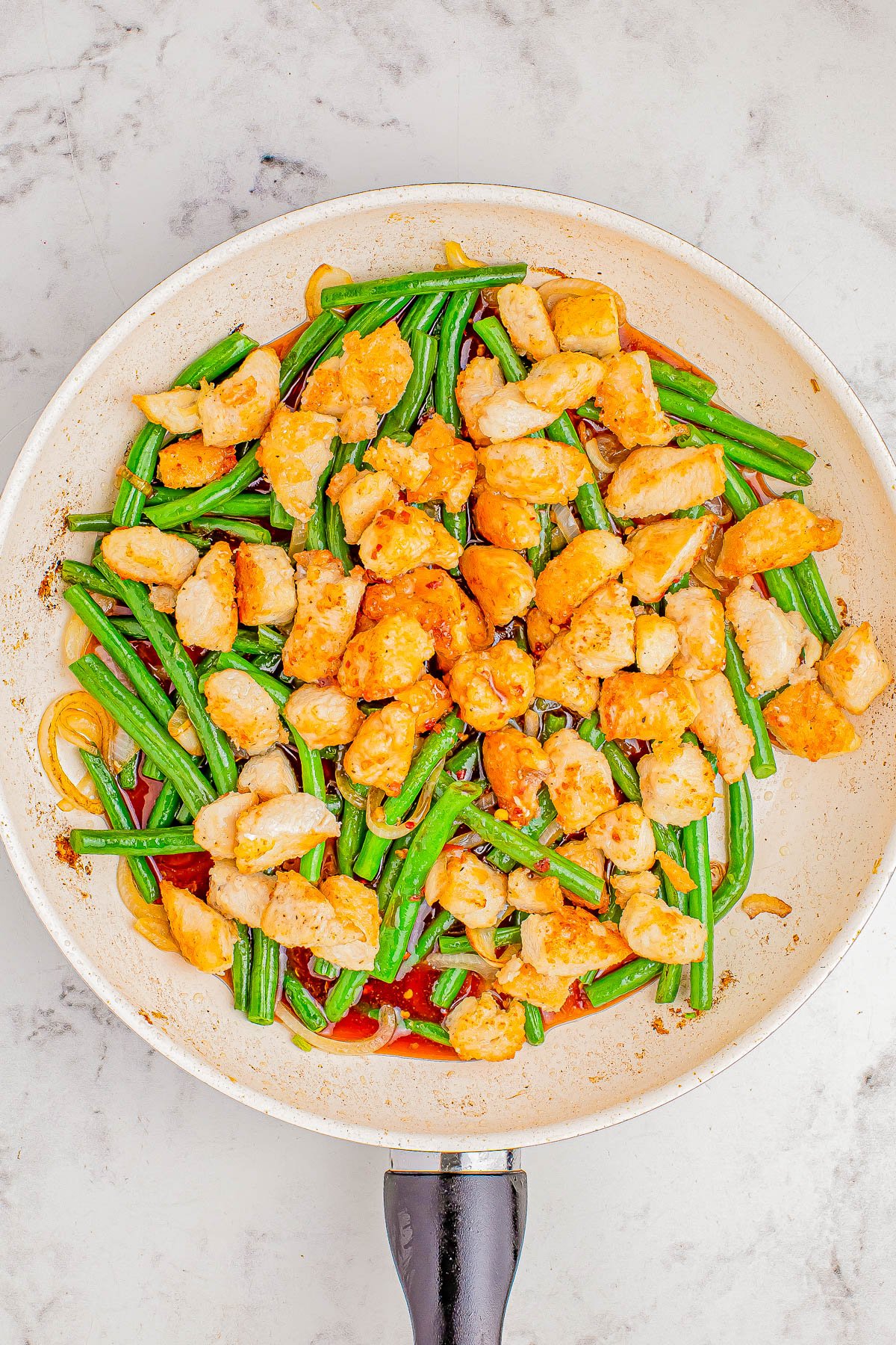 A stir-fried dish with chicken, green beans, and red peppers in a skillet, on a marble countertop.