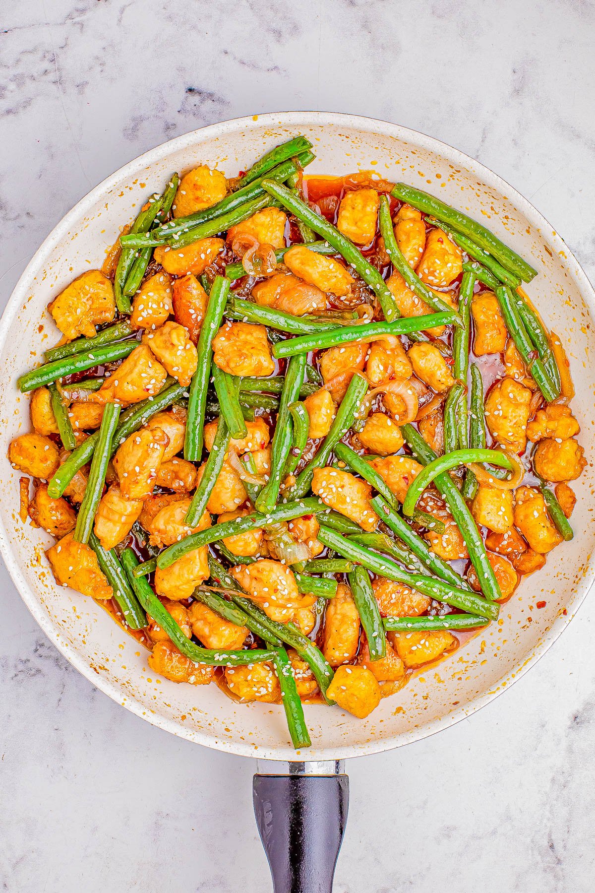 Stir-fried chicken and green beans in a sauce, served in a skillet on a marble surface.