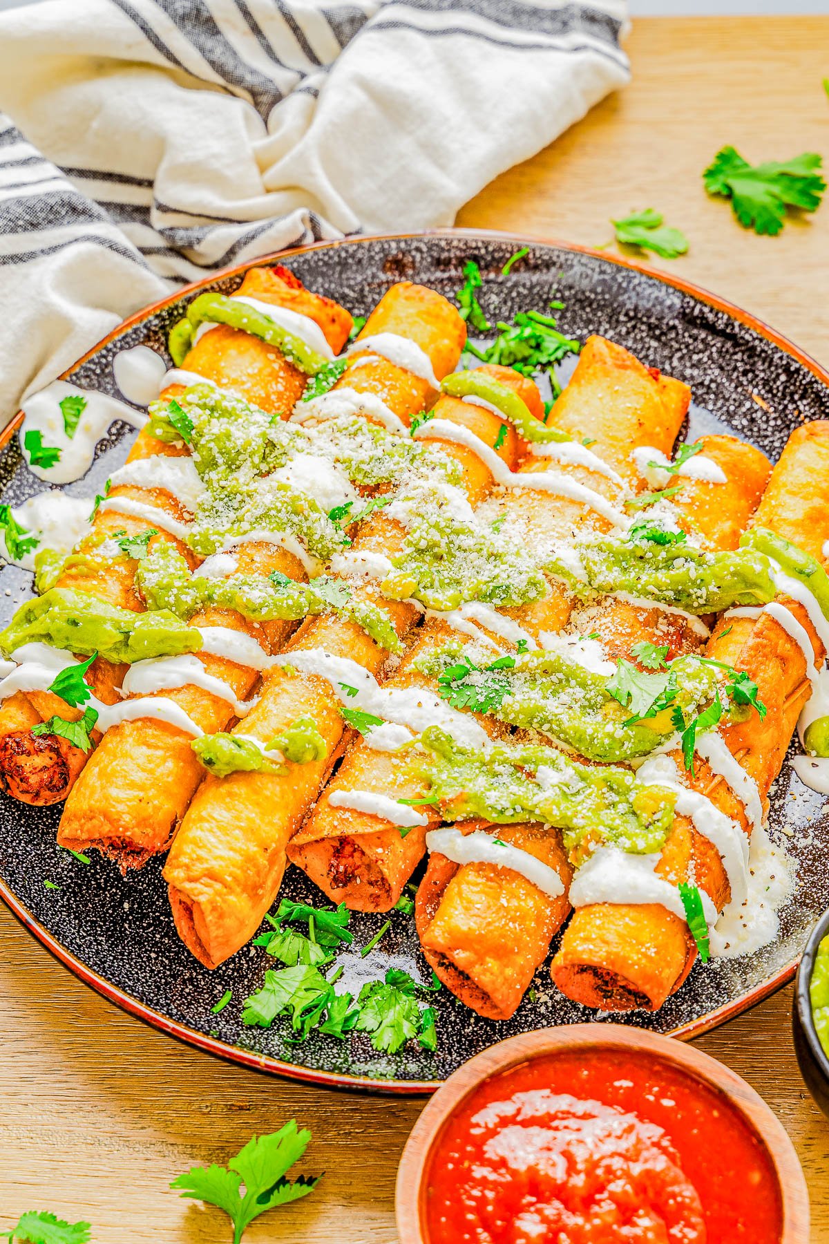 Plate of crispy taquitos drizzled with green sauce and sour cream, garnished with cilantro, served with a side of red salsa.