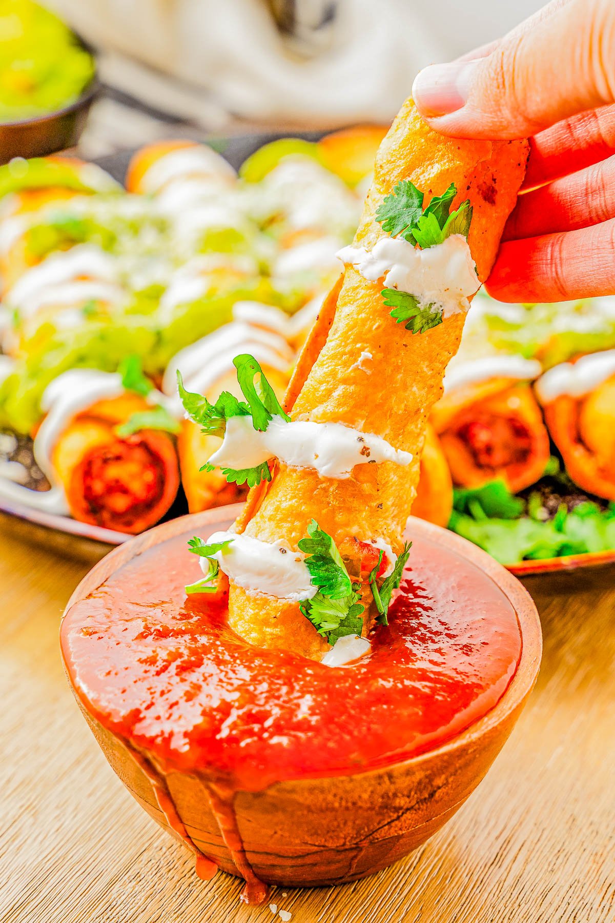 A person dipping a taquito into a bowl of red salsa, garnished with sour cream and cilantro, with more taquitos in the background.