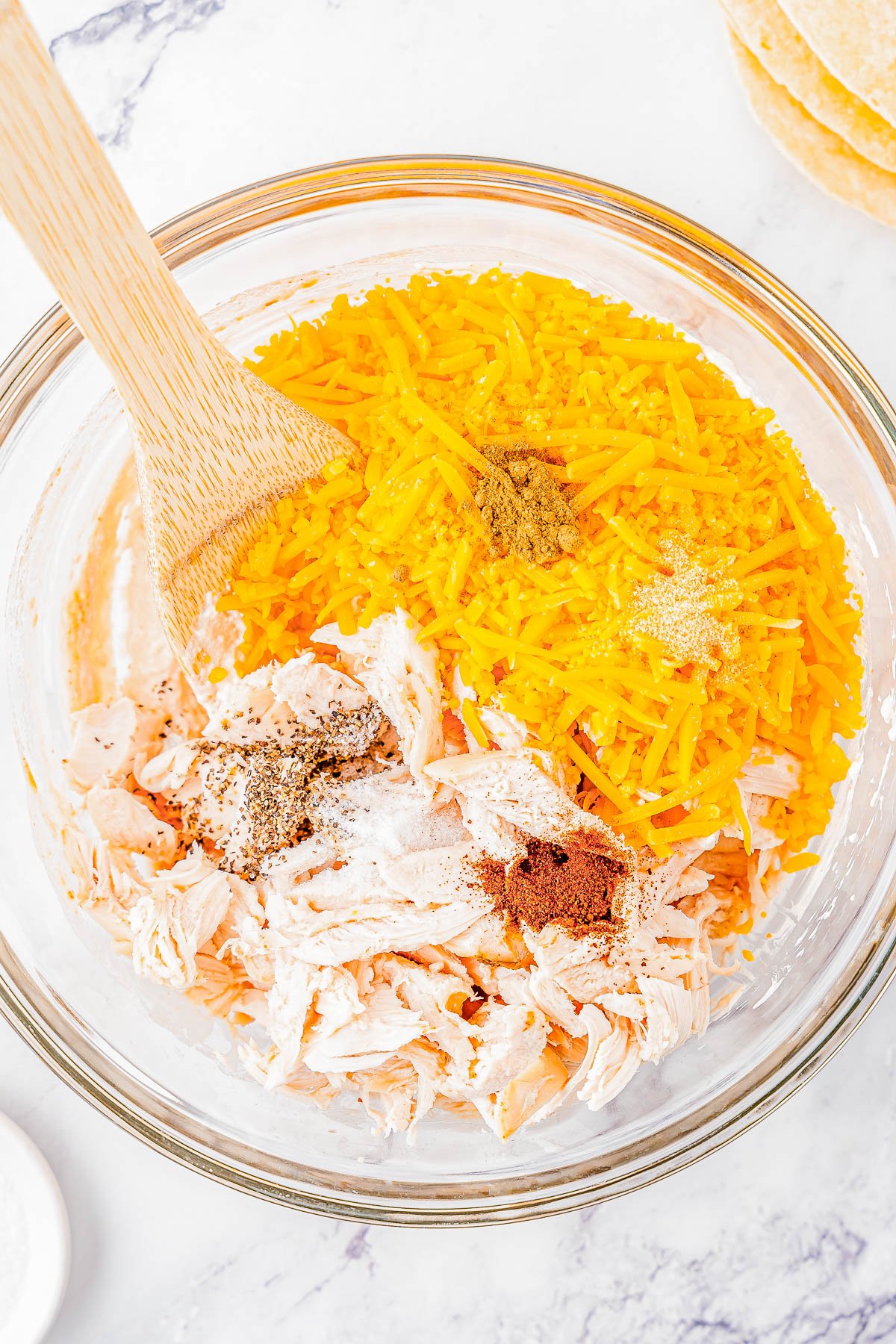 Glass bowl of shredded chicken and seasoned with spices, mixed with a wooden spoon, on a marble surface.