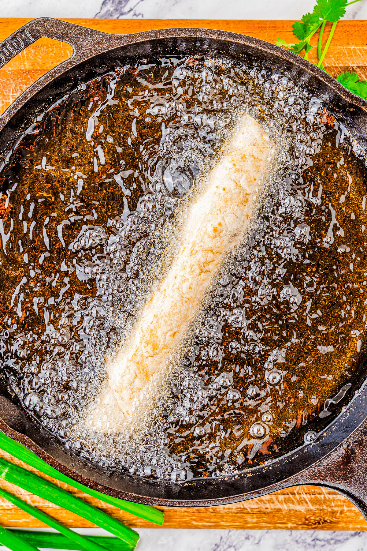 Taquito frying in a cast iron skillet, surrounded by bubbling oil, with fresh herbs and green onions nearby.