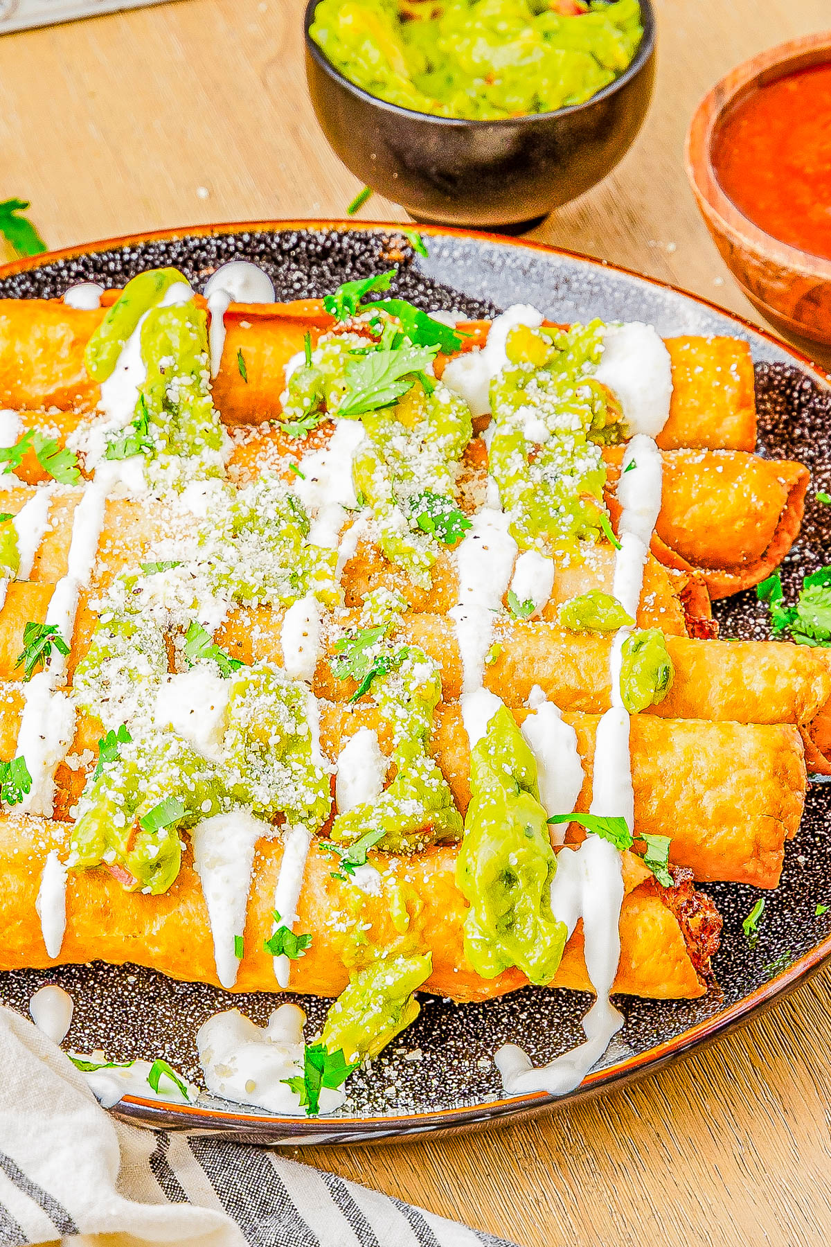 A plate of crispy taquitos topped with green salsa, sour cream, and crumbled cheese, garnished with fresh cilantro.