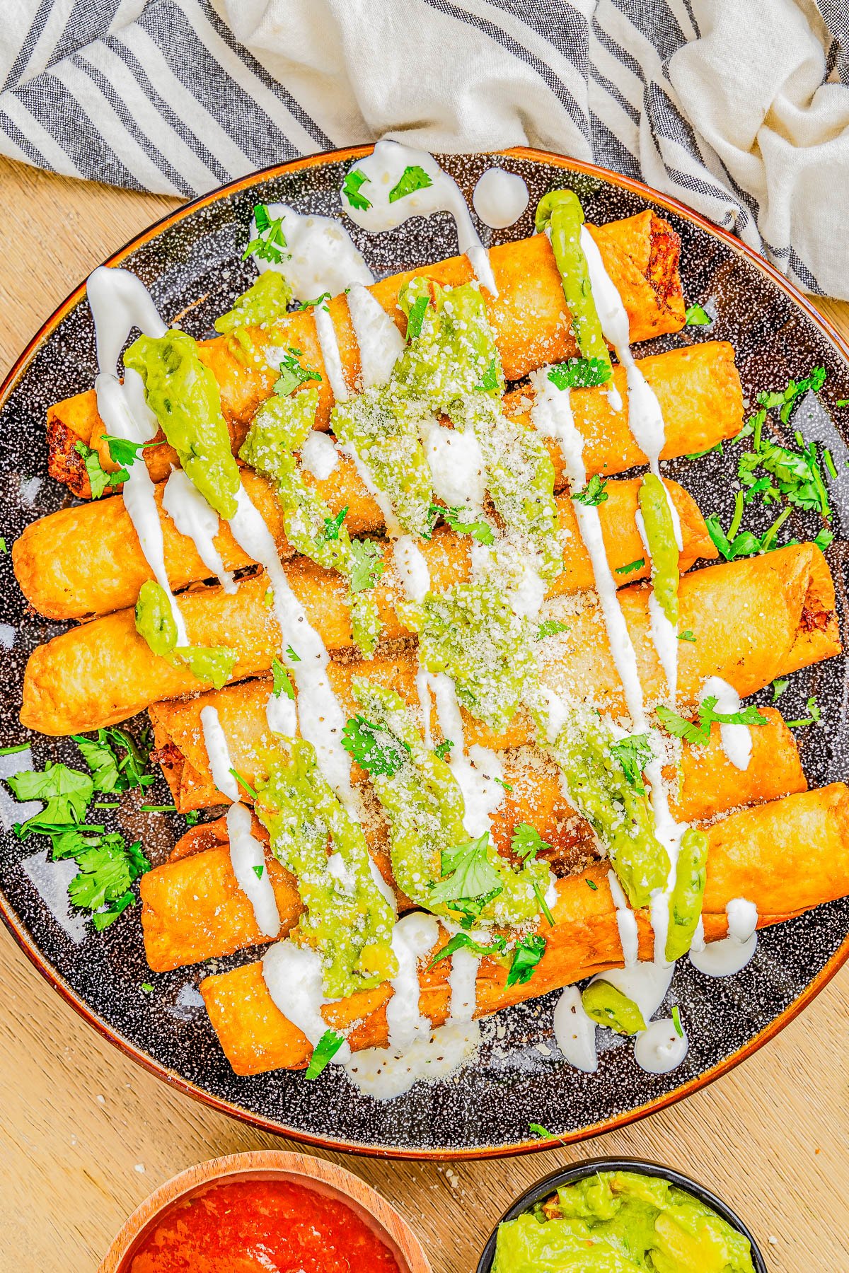 A plate of taquitos covered in green and white sauces, sprinkled with cheese, served with sides of red and green salsas and guacamole.