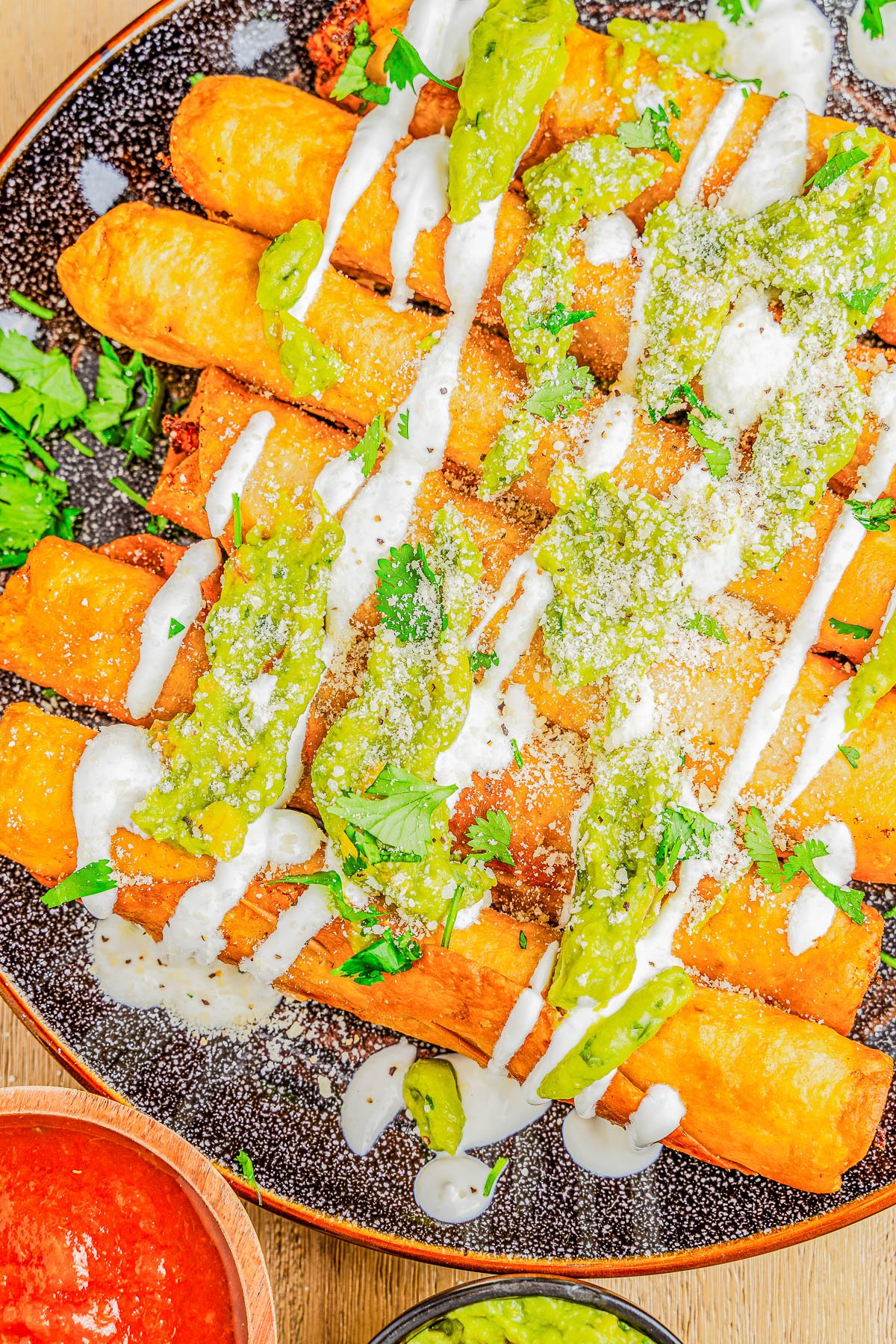 Plate of golden-brown taquitos drizzled with green sauce and cream, garnished with grated cheese and cilantro, served with a side of red salsa.