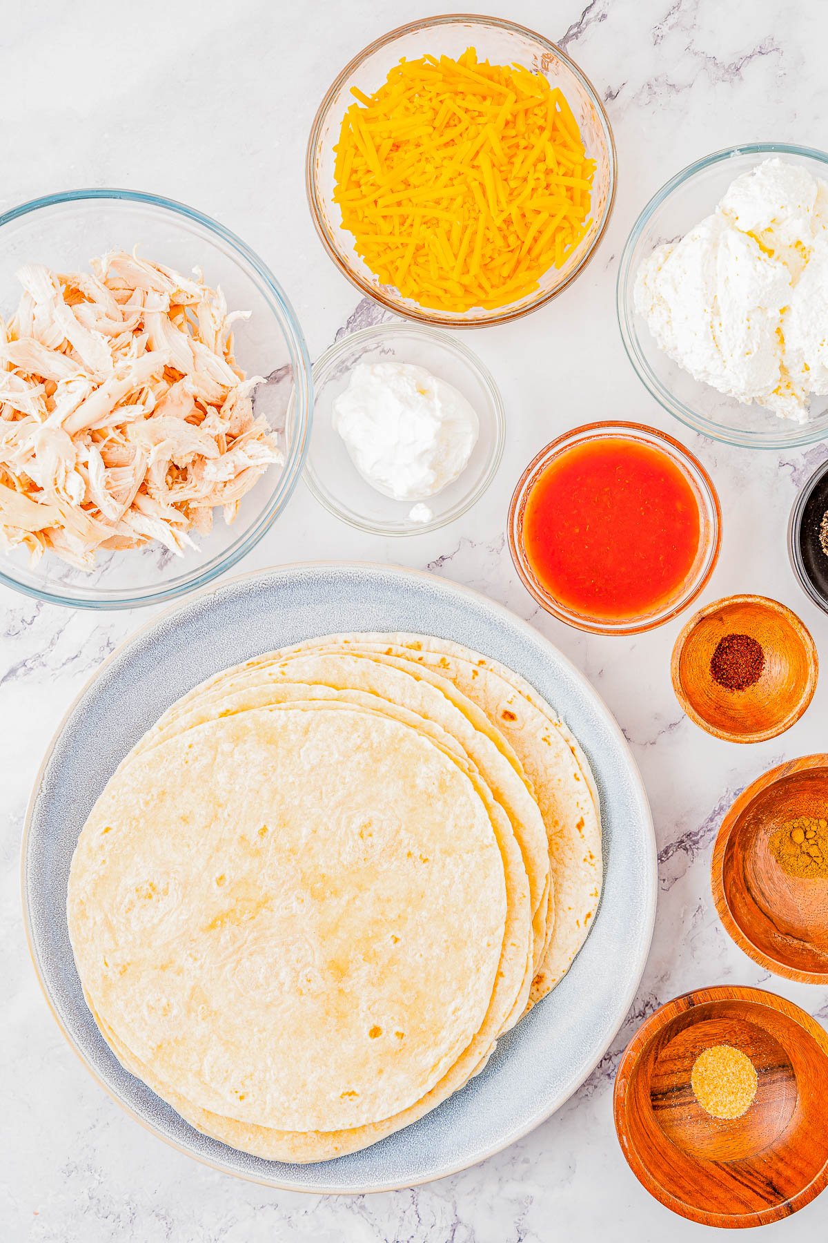 Ingredients for making taquitos, including shredded chicken, cheddar cheese, sour cream, salsa, and tortillas, displayed in separate bowls on a marble countertop.