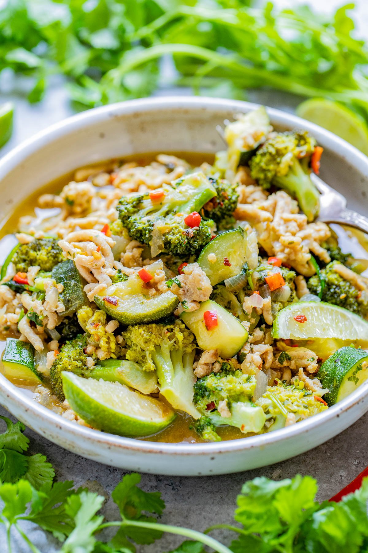 A bowl of green curry with ground chicken, broccoli, zucchini, and herbs, garnished with red chili slices.