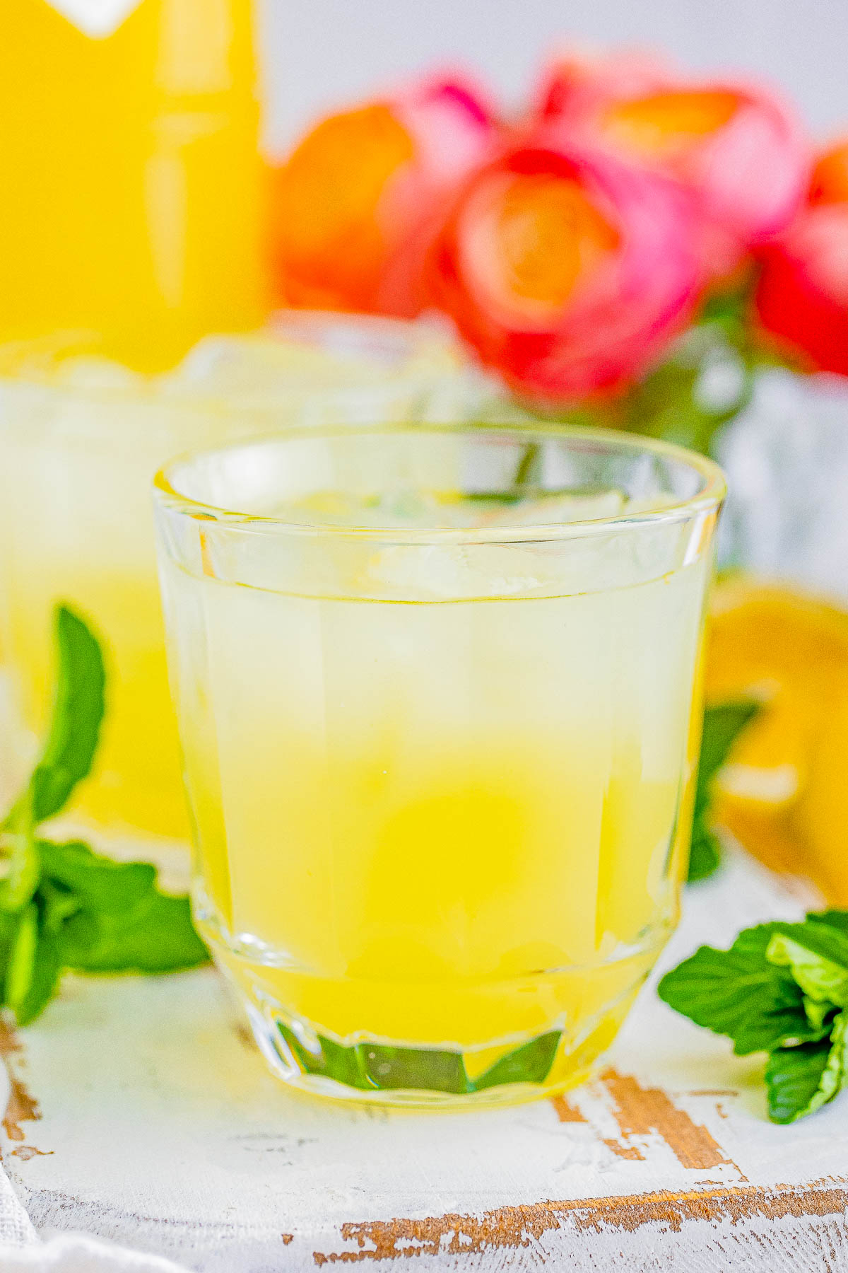 A glass of limoncello with ice cubes, garnished with mint leaves, accompanied by fresh lemons and flowers in the background.