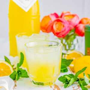 A refreshing limoncello spritz with mint leaves, surrounded by sliced lemons, a bottle of lemon juice, and vibrant flowers in the background.