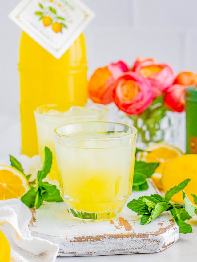 A refreshing limoncello spritz with mint leaves, surrounded by sliced lemons, a bottle of lemon juice, and vibrant flowers in the background.