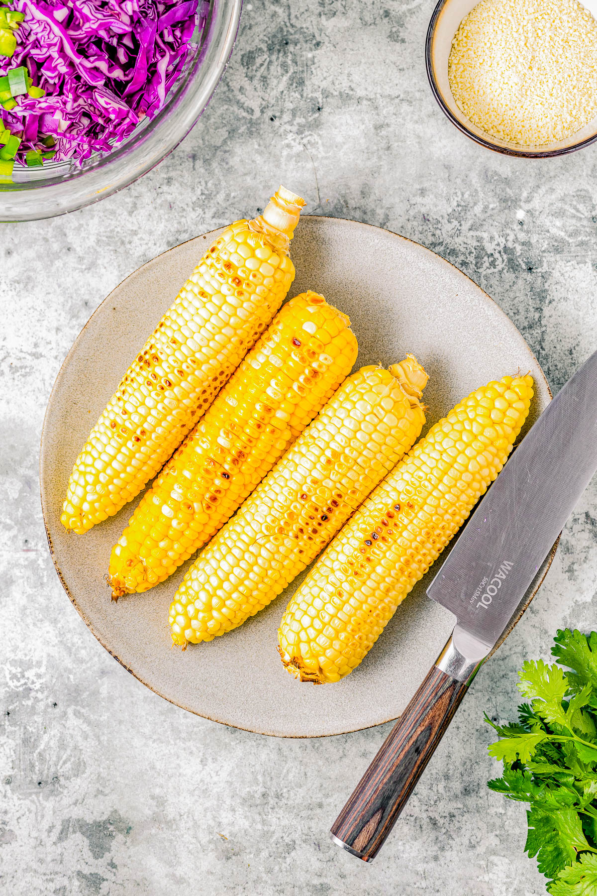 Four grilled corn cobs on a plate with a knife on the side and bowls of sesame seeds and shredded purple cabbage in the background.
