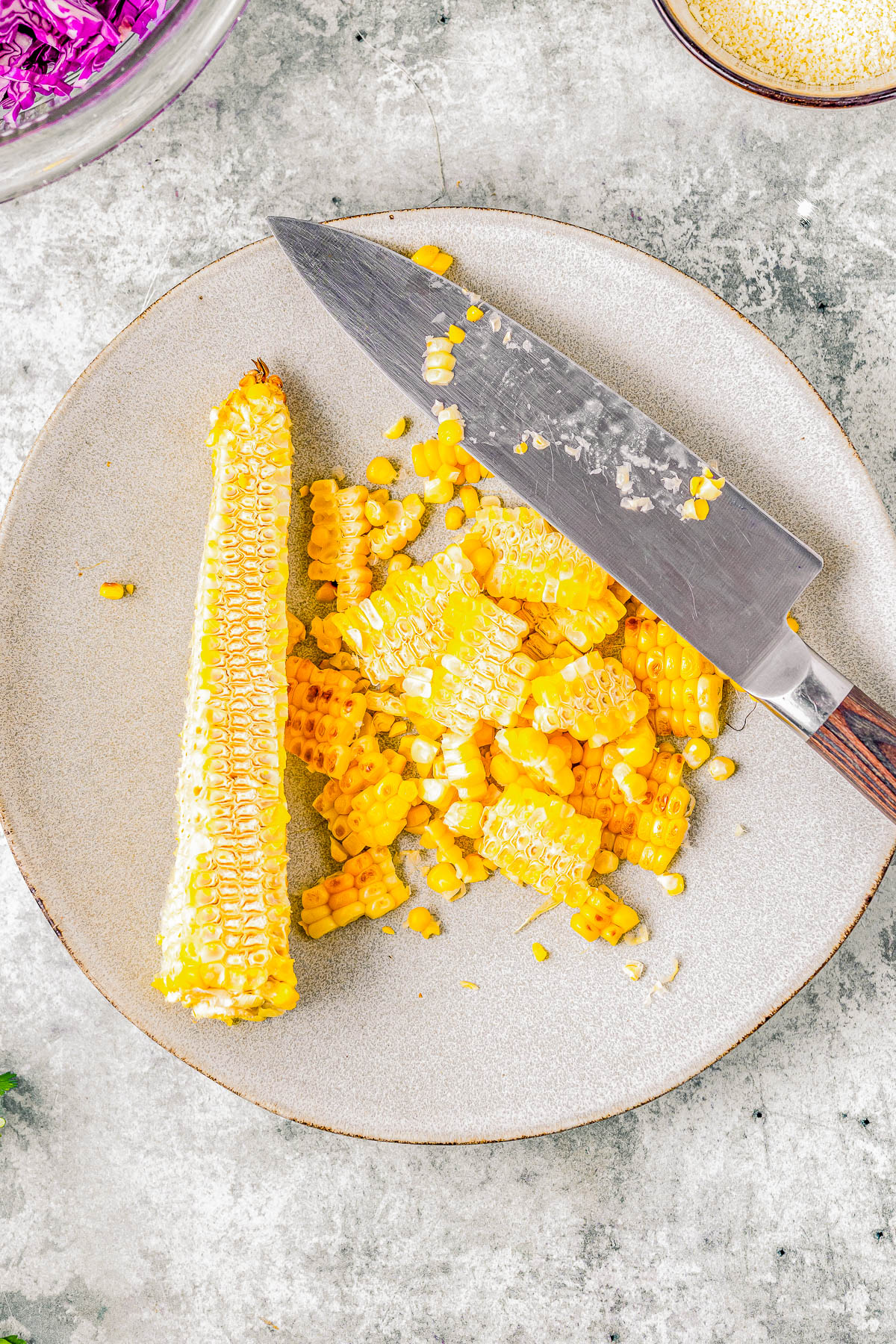 A knife cutting through corn on the cob on a plate, with kernels scattered and a bowl of cabbage nearby on a textured surface.