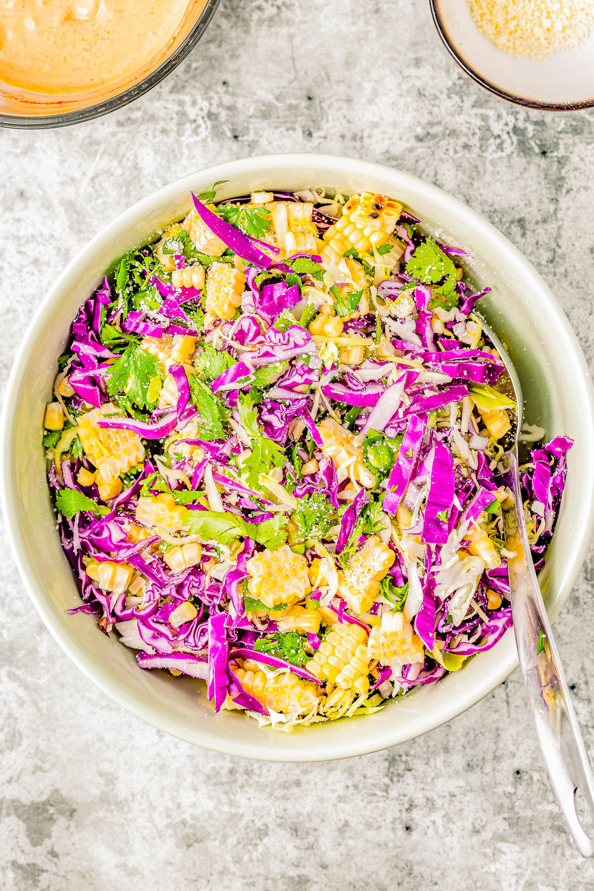 A bowl of vibrant salad containing red cabbage, carrots, herbs, and sesame seeds, with a spoon on the side and dressing in the background.