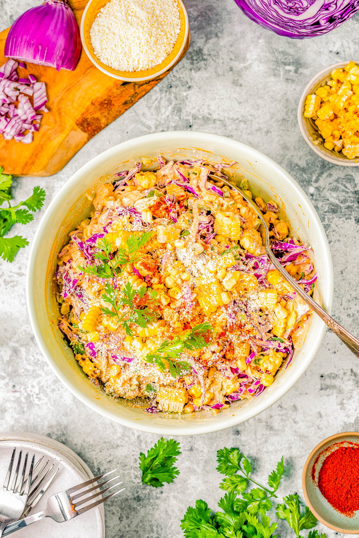 A vibrant bowl of corn salad with purple cabbage and parsley, surrounded by bowls of ingredients and spices on a textured surface.