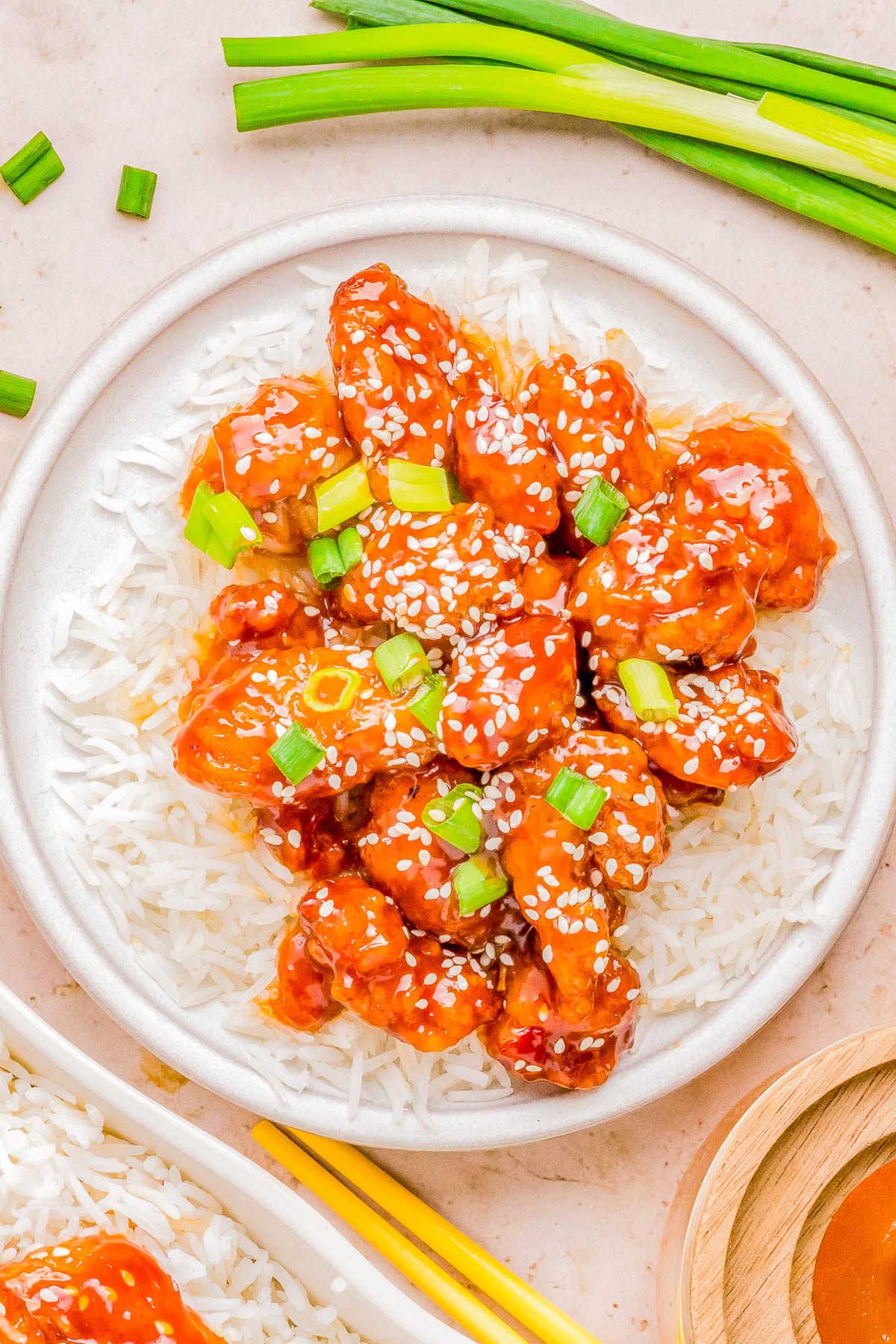 A plate of sesame chicken served over white rice, garnished with green onions and sesame seeds, with chopsticks on the side.