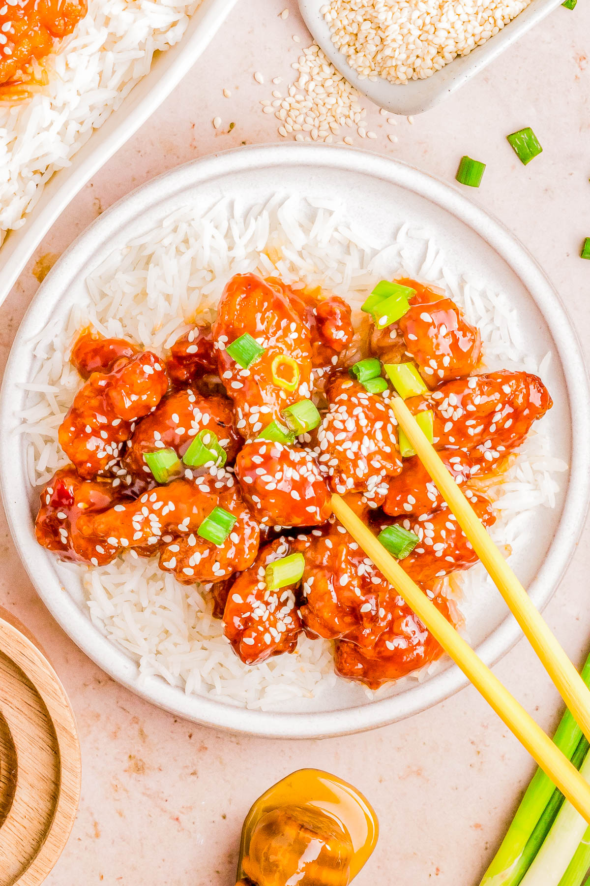 A plate of sesame chicken garnished with sesame seeds and green onions, served over white rice, with chopsticks on the side.