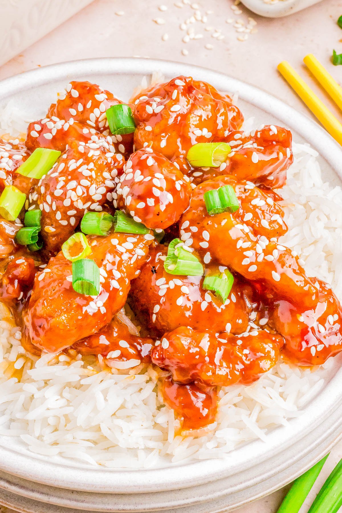 A plate of sesame chicken served over white rice, garnished with green onions and sesame seeds, with chopsticks on the side.