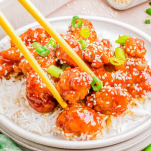 Plate of sesame chicken over rice with chopsticks, garnished with sesame seeds and green onions on a light tabletop.