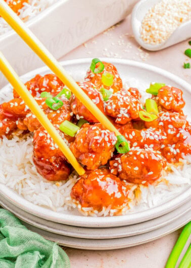 Plate of sesame chicken over rice with chopsticks, garnished with sesame seeds and green onions on a light tabletop.