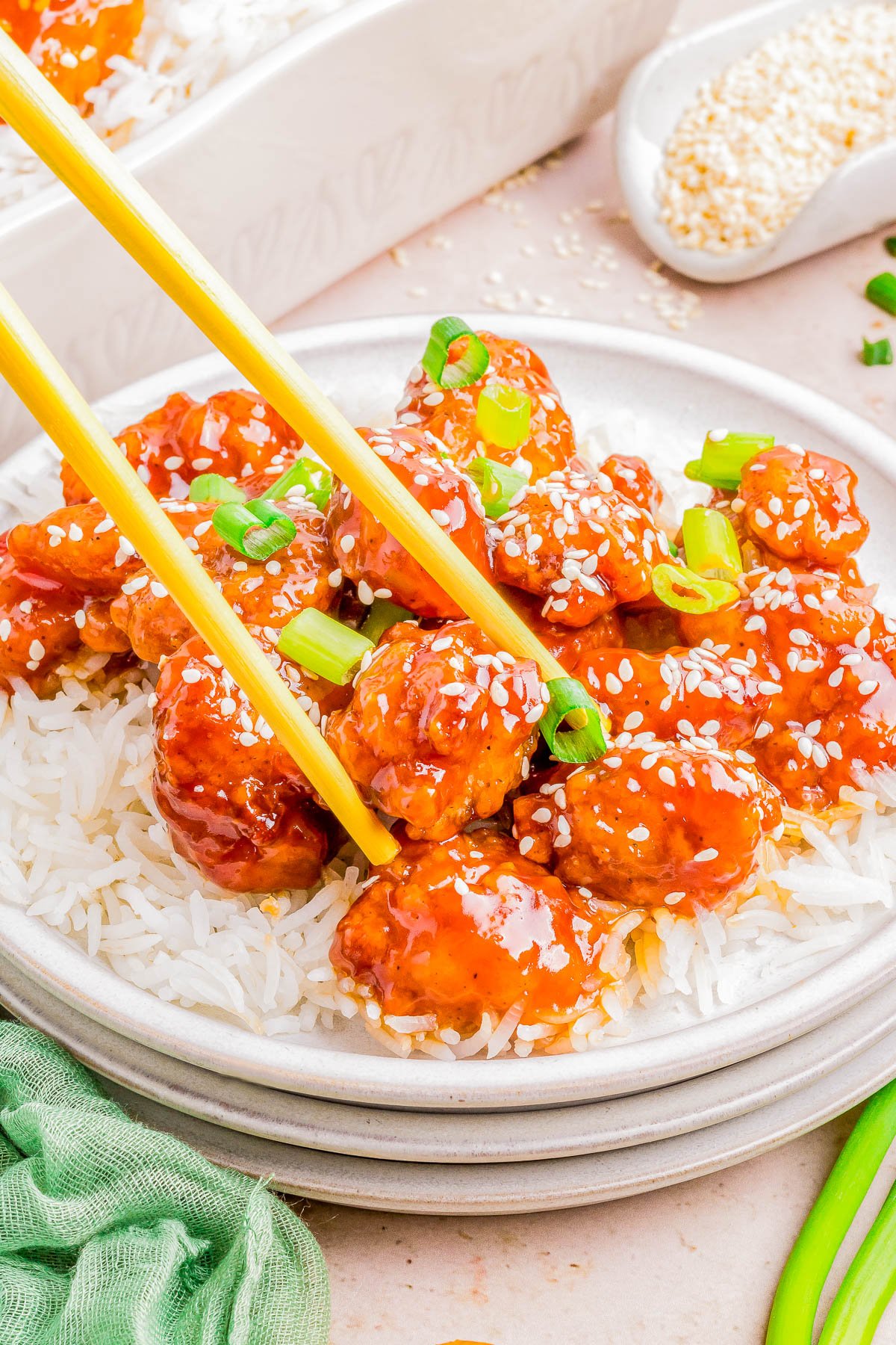 Plate of sesame chicken over rice with chopsticks, garnished with sesame seeds and green onions on a light tabletop.