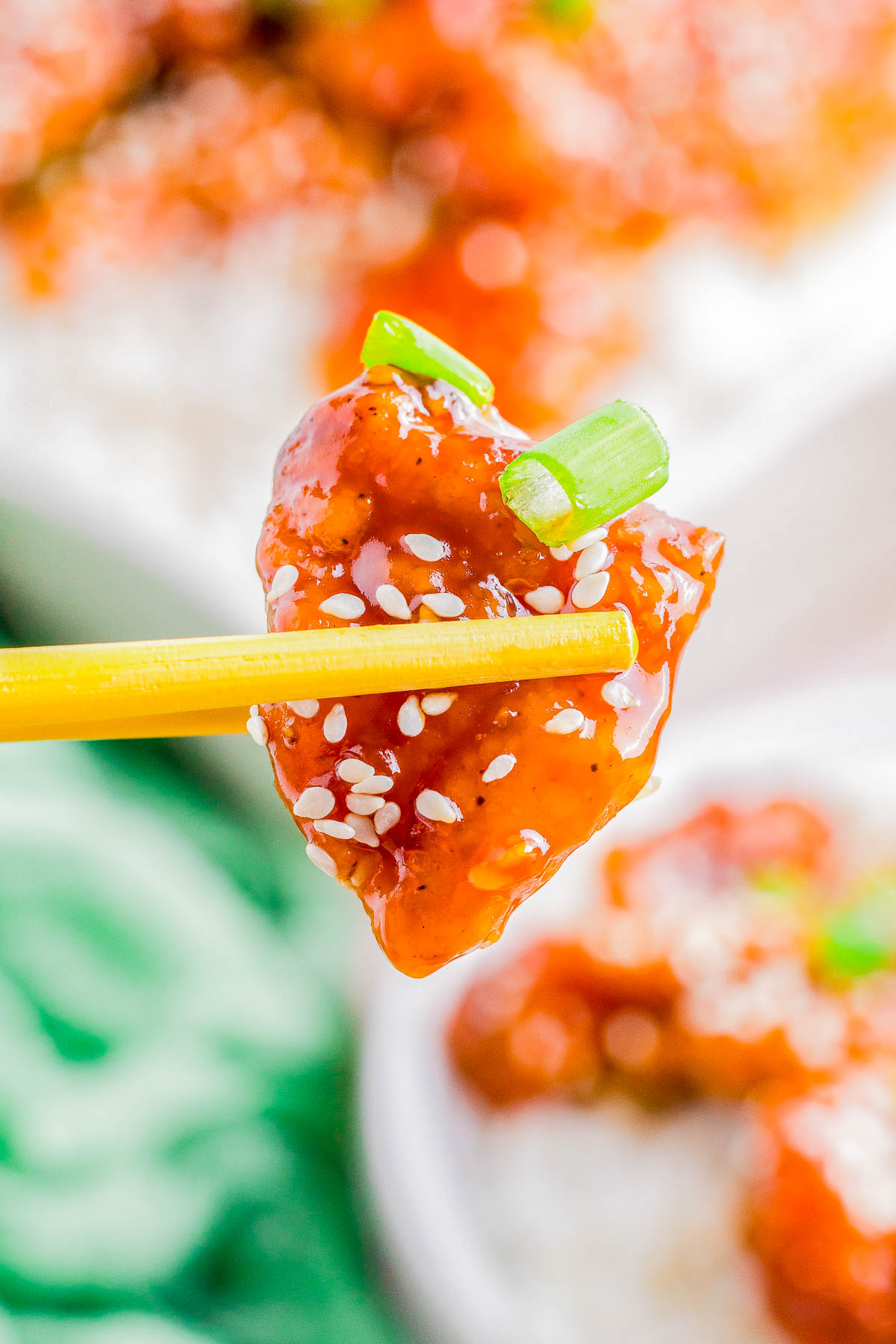 Chopsticks holding a piece of sesame chicken coated in a glossy red sauce, sprinkled with sesame seeds and garnished with green onions.