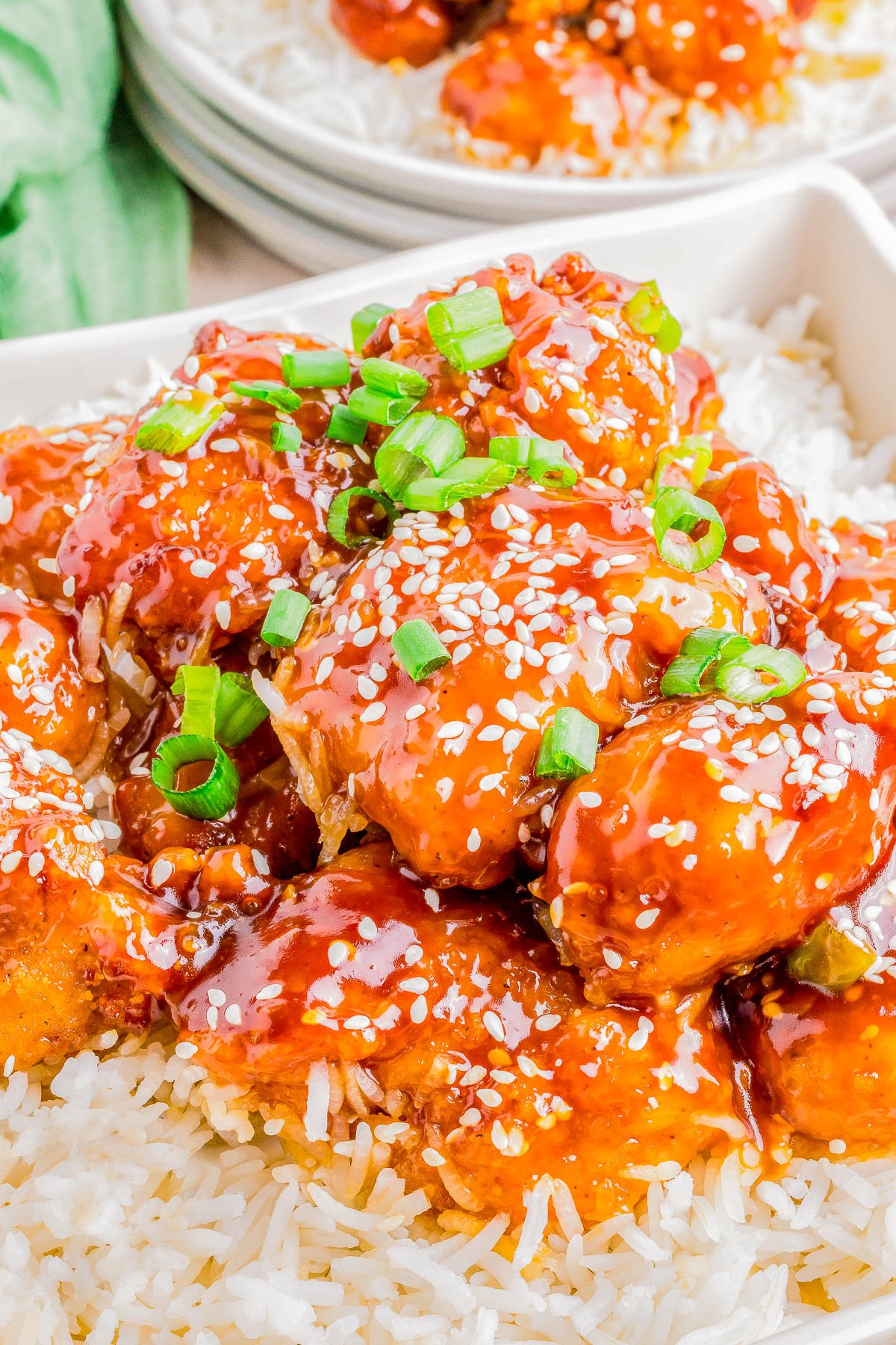 A plate of sesame chicken topped with scallions served over white rice, garnished with sesame seeds.