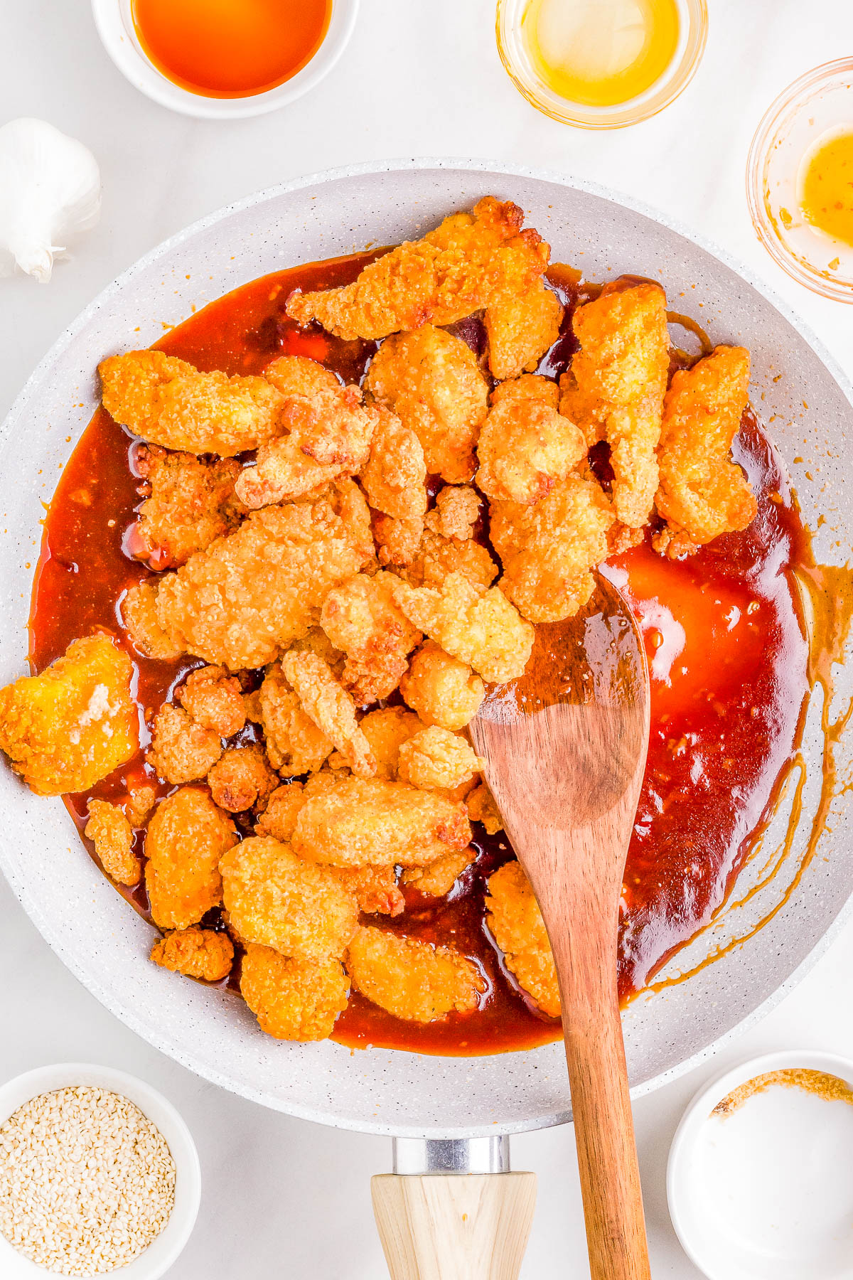 Baked chicken pieces being tossed in a sauce in a white skillet, surrounded by ingredients like honey and garlic.