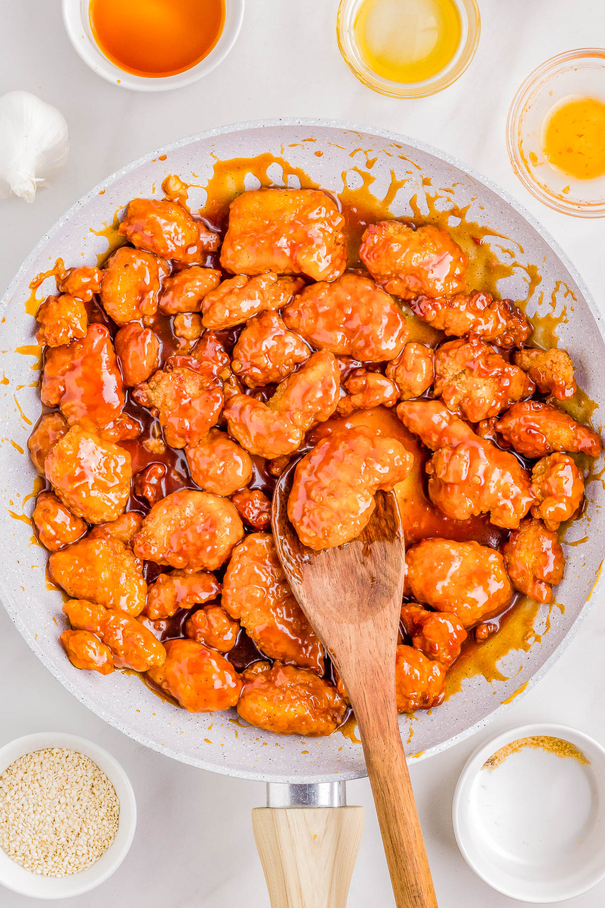 Sautéed sesame chicken in a pan, coated in sticky orange sauce, with a wooden spoon, surrounded by small bowls of ingredients and sesame seeds.