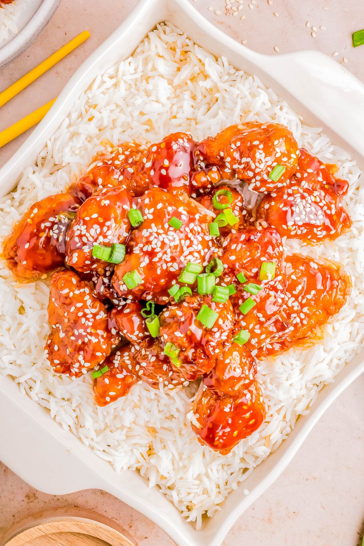 A dish of sesame chicken over white rice, garnished with scallions and sesame seeds, served on a white plate with chopsticks on the side.