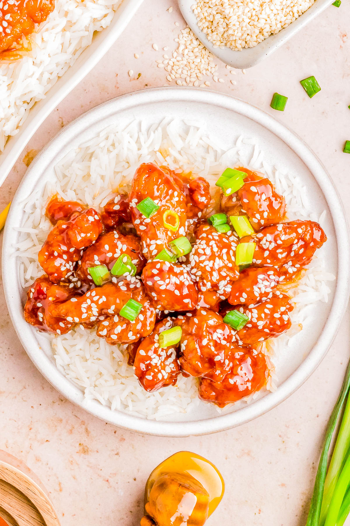 A plate of sesame chicken over white rice garnished with sesame seeds and green onions, with sesame seeds and green onions on the side.