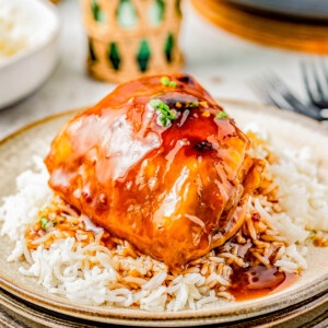 A plate of shoyu chicken served over steamed rice, garnished with green onions, presented on a rustic plate with cutlery and a woven basket in the background.