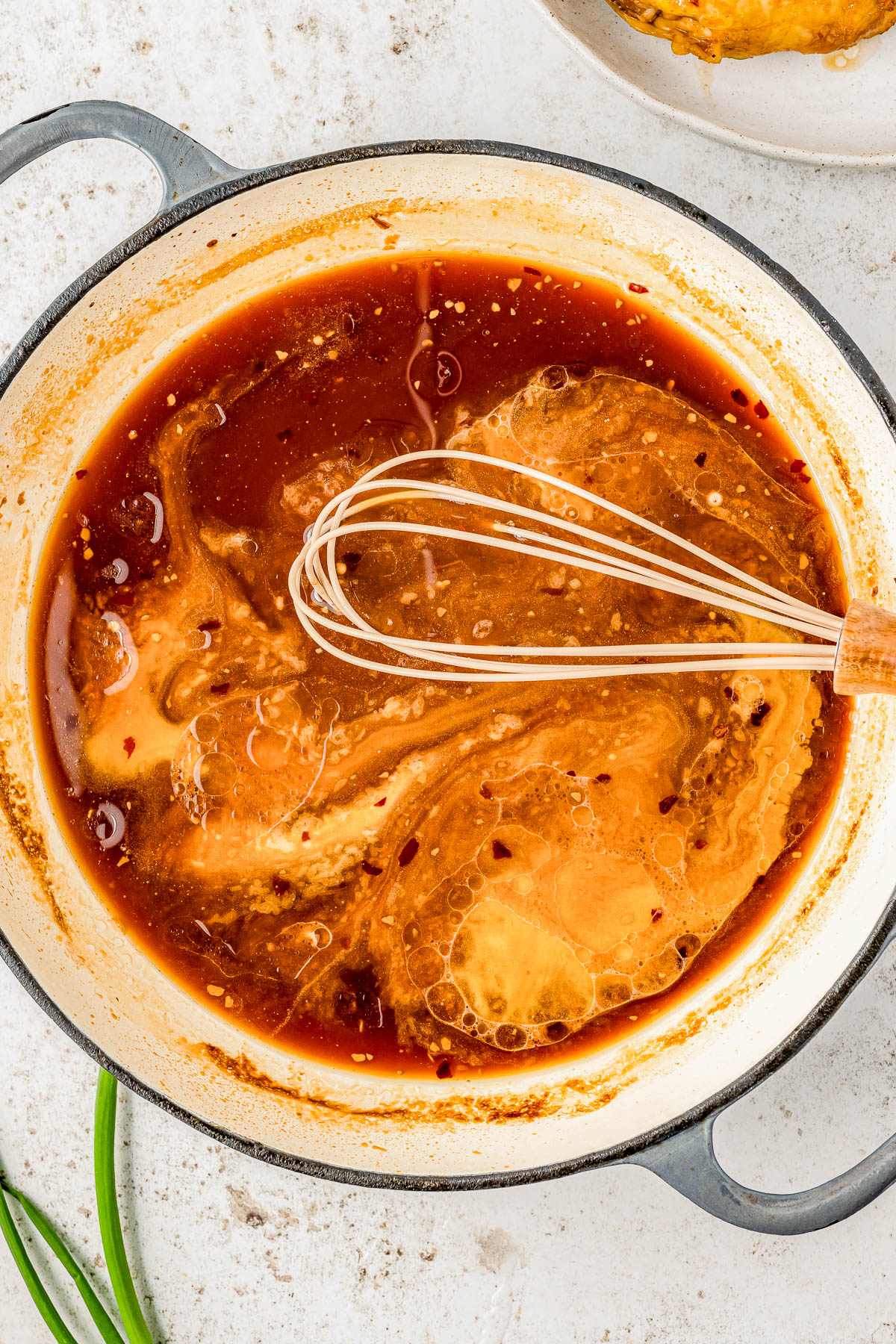 Close-up of a whisk stirring a rich, bubbling red sauce in a black pot, with hints of spices and oil droplets on the surface.