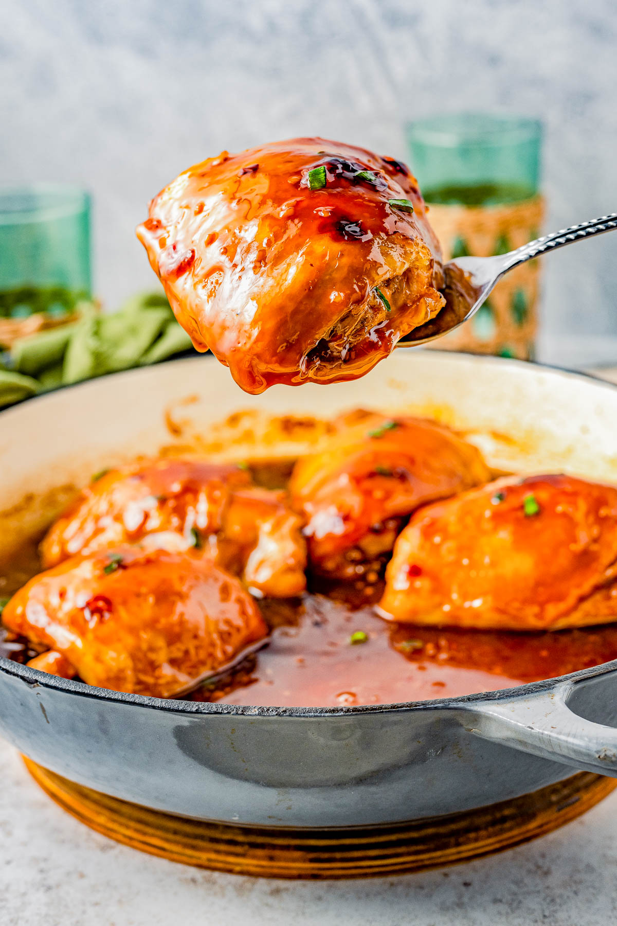 A spoon lifting a glazed chicken thigh from a skillet, with more chicken and sauce in the background, garnished with herbs.