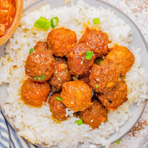 A bowl of white rice topped with glazed meatballs, garnished with sesame seeds and green onions.