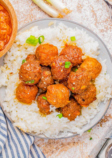 A bowl of white rice topped with glazed meatballs, garnished with sesame seeds and green onions.