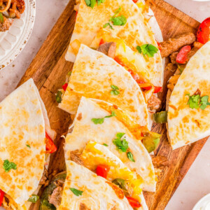 Platter of steak quesadillas garnished with peppers, tomatoes, and cilantro, served with lime wedges and dipping sauces on a wooden board.