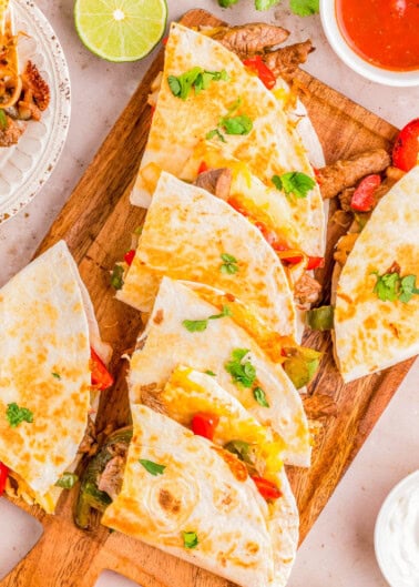 Platter of steak quesadillas garnished with peppers, tomatoes, and cilantro, served with lime wedges and dipping sauces on a wooden board.