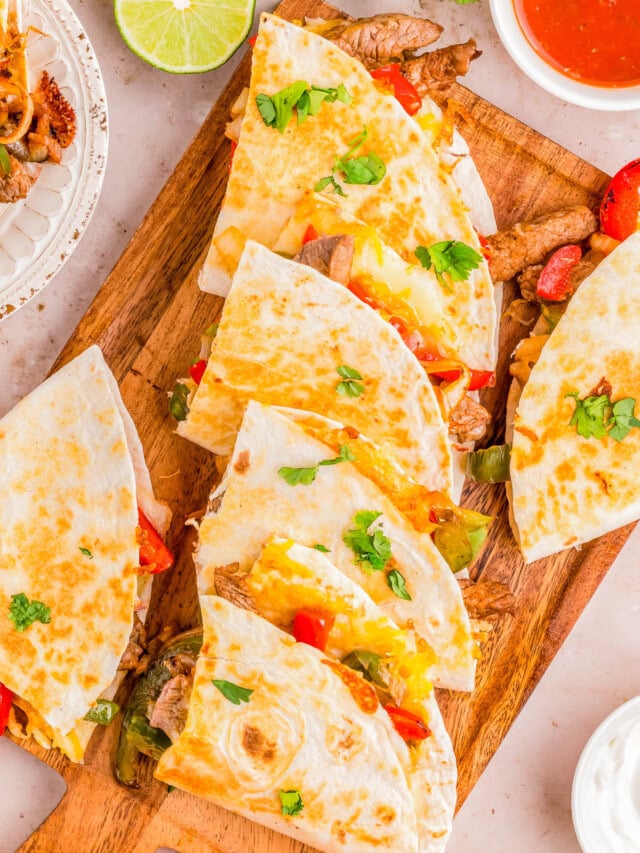 Platter of steak quesadillas garnished with peppers, tomatoes, and cilantro, served with lime wedges and dipping sauces on a wooden board.