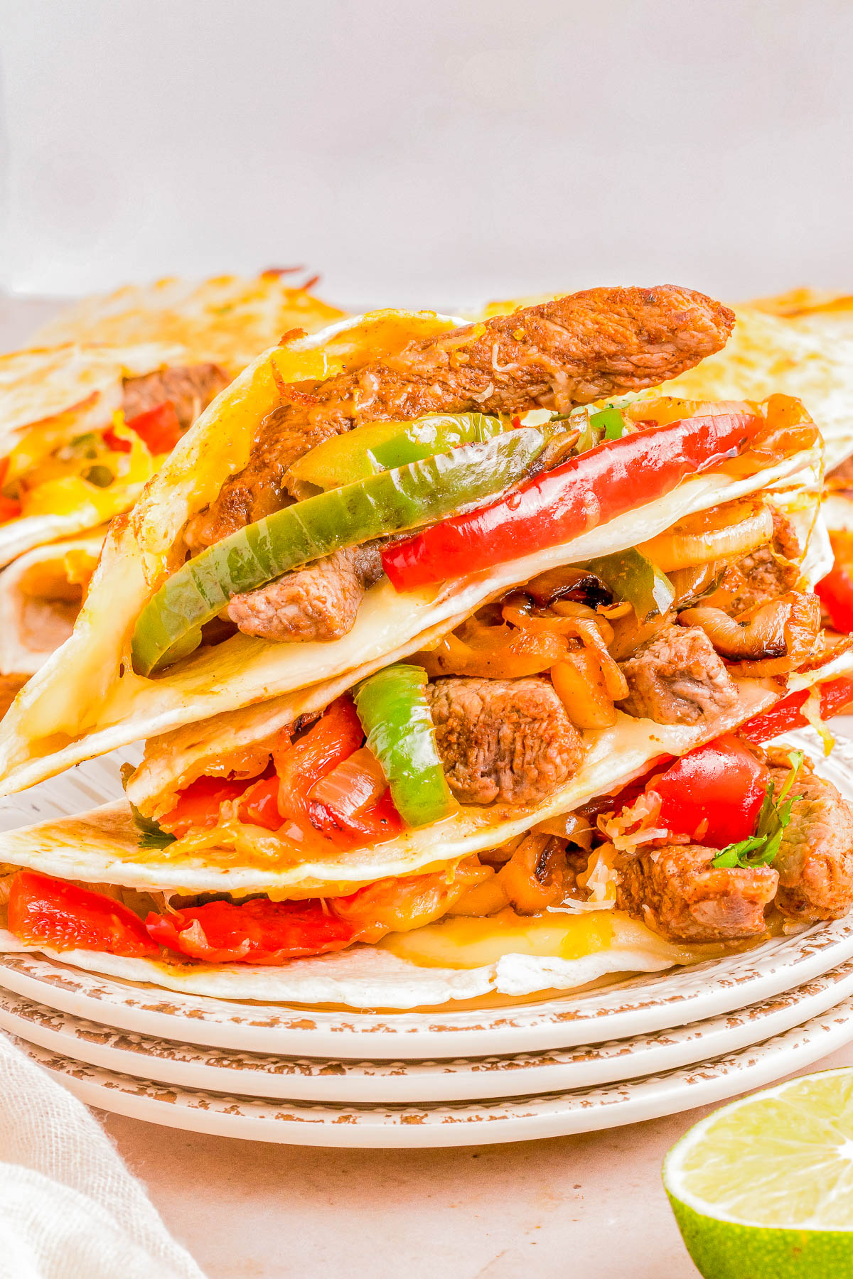 A stack of steak quesadillas with melted cheese, bell peppers, onions, on a plate, garnished with lime slices.