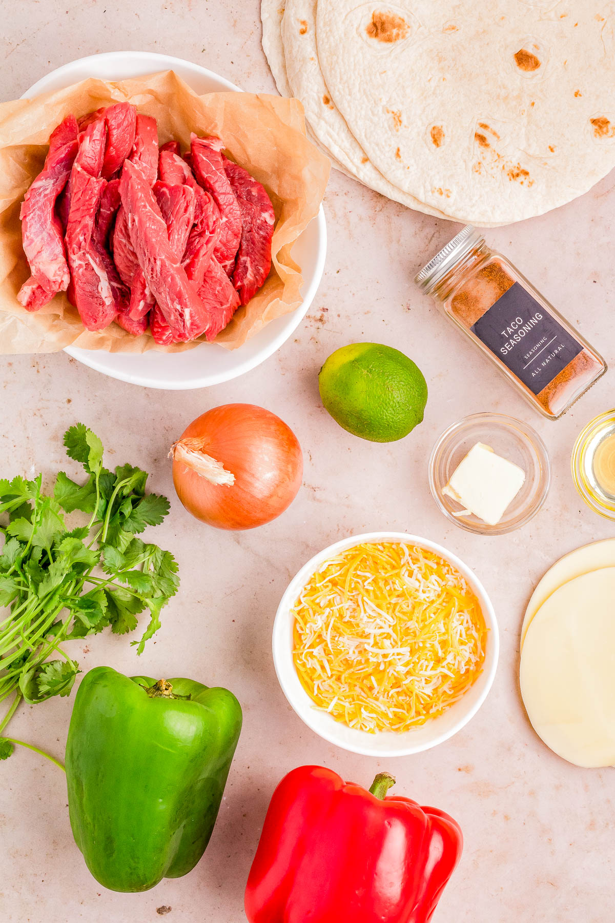 Ingredients for cooking displayed on a table, including raw beef strips, tortillas, cheese, lime, bell peppers, onion, cilantro, and spices.
