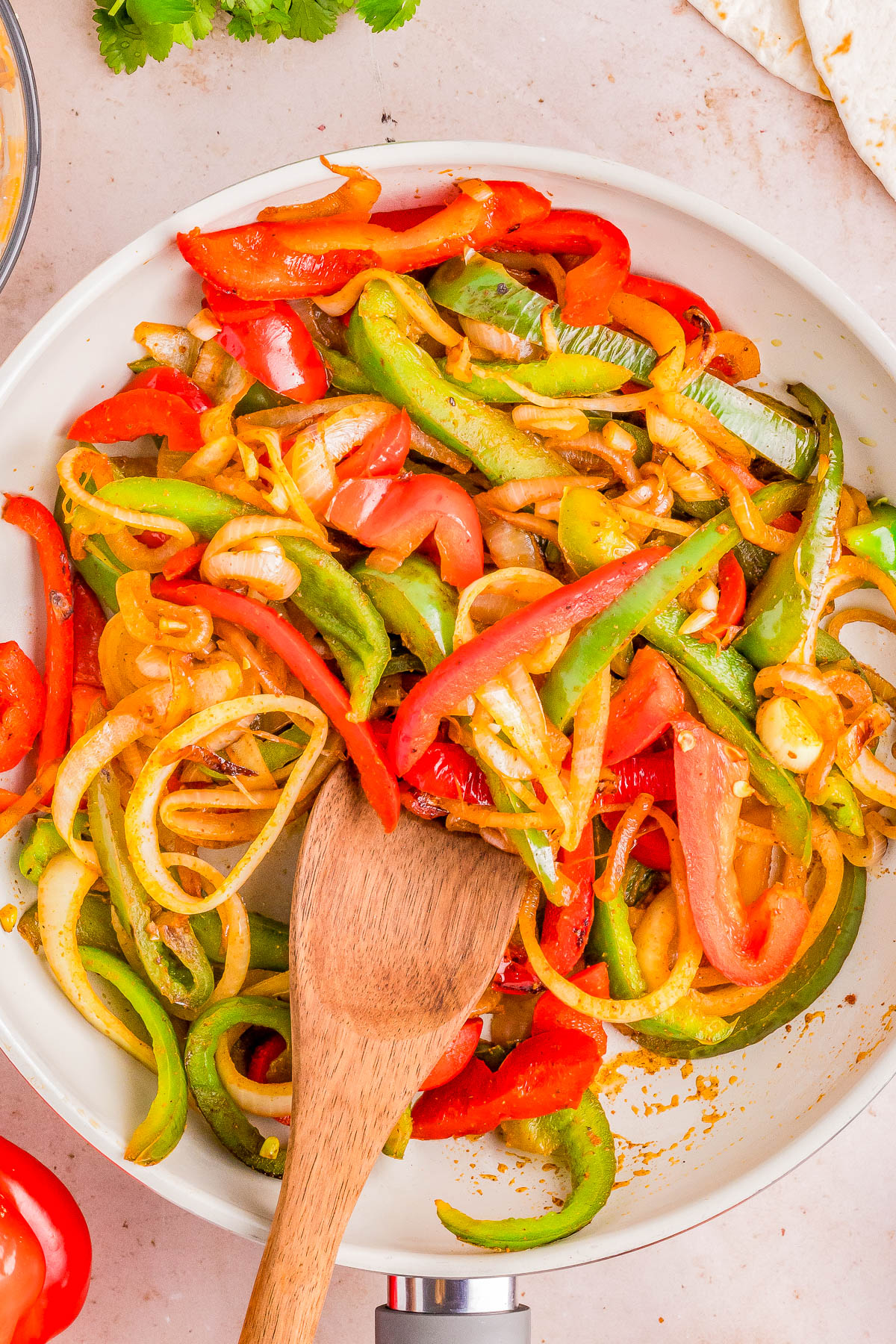 A pan filled with sautéed red, green, and yellow bell peppers and onions, mixed with a wooden spoon.